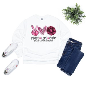 Może przedstawiać: Biała koszulka z długim rękawem z r&oacute;żowym znakiem pokoju, sercem i kwiatem. Projekt zawiera słowa "Peace Love Cure" i "Breast Cancer Awareness". Koszulka jest połączona z niebieskimi dżinsami i białymi trampkami.