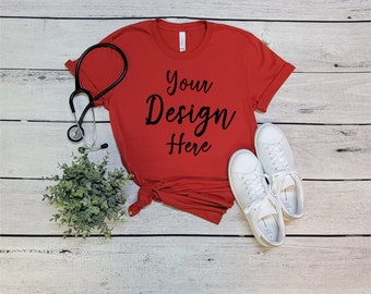 May include: Red t-shirt with the text "Your Design Here" on a white wood background. A stethoscope and a pair of white sneakers are also in the image.