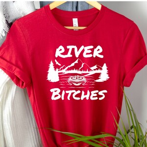 Pu&ograve; includere: Maglietta rossa con testo bianco che dice "River Bitches" e una grafica di una scena di fiume con alberi e montagne.