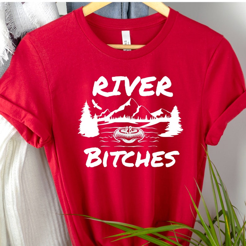 Pu&ograve; includere: Maglietta rossa con testo bianco che dice "River Bitches" e una grafica di una scena di fiume con alberi e montagne.