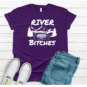 Pu&ograve; includere: Maglietta viola con testo bianco che recita "River Bitches" e una grafica di un lago con montagne e alberi.