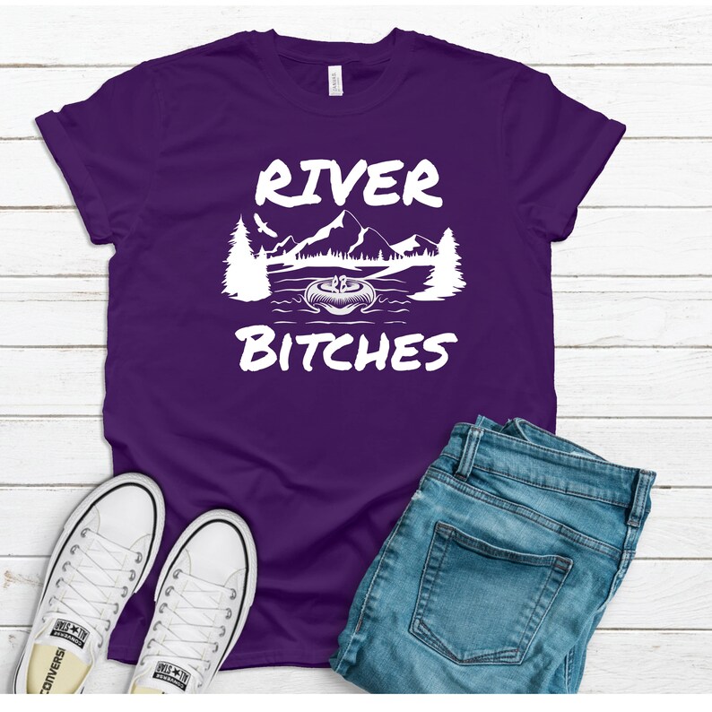 Pu&ograve; includere: Maglietta viola con testo bianco che recita "River Bitches" e una grafica di un lago con montagne e alberi.