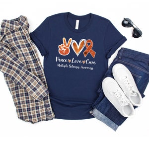 Puede incluir: Camiseta azul marino con un signo de paz, un coraz&oacute;n y una cinta naranja con el texto "Peace Love Cure" y "Multiple Sclerosis Awareness".