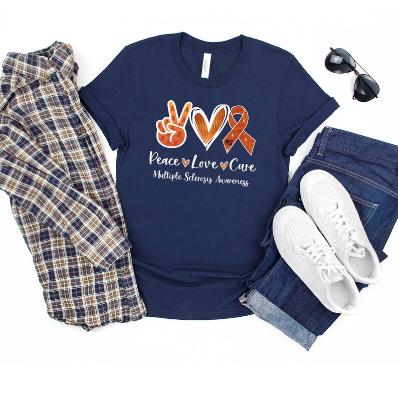 Puede incluir: Camiseta azul marino con un signo de paz, un coraz&oacute;n y una cinta naranja con el texto "Peace Love Cure" y "Multiple Sclerosis Awareness".