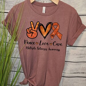 Puede incluir: Una camiseta color malva con un signo de paz, un coraz&oacute;n y una cinta naranja con el texto "MS" en su interior. El texto debajo dice "Peace Love Cure Multiple Sclerosis Awareness".