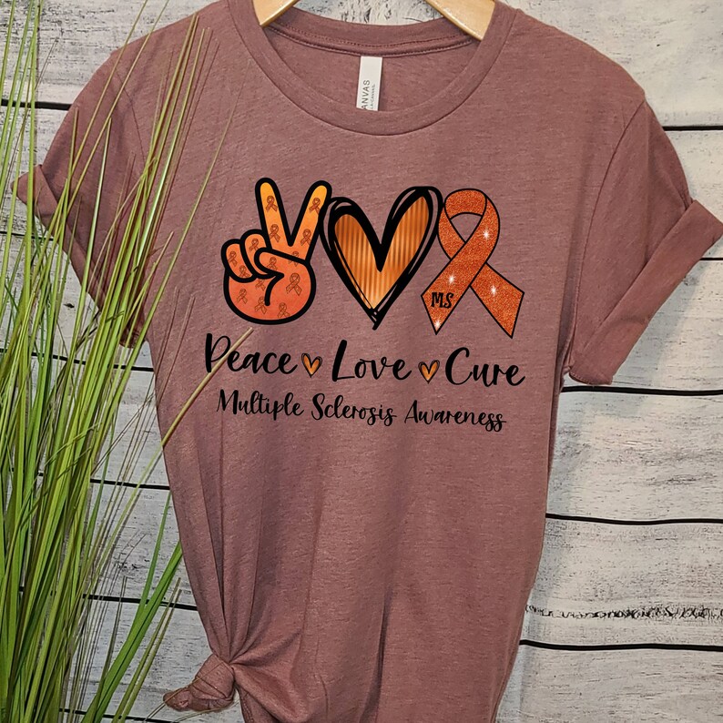 Puede incluir: Una camiseta color malva con un signo de paz, un coraz&oacute;n y una cinta naranja con el texto "MS" en su interior. El texto debajo dice "Peace Love Cure Multiple Sclerosis Awareness".