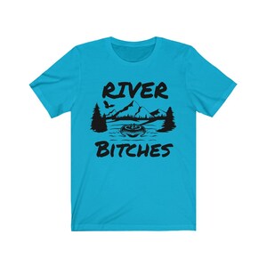 May include: A turquoise blue t-shirt with a black graphic design of a river scene with mountains, trees, and a boat. The text "River Bitches" is printed on the shirt.