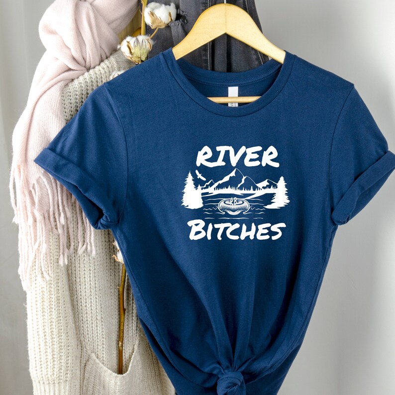 Pu&ograve; includere: Una maglietta blu navy con una grafica bianca di una scena di montagna e il testo "River Bitches".