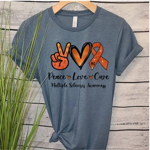 Puede incluir: Una camiseta azul con un signo de paz, un coraz&oacute;n y una cinta naranja con el texto "Peace Love Cure" y "Multiple Sclerosis Awareness".