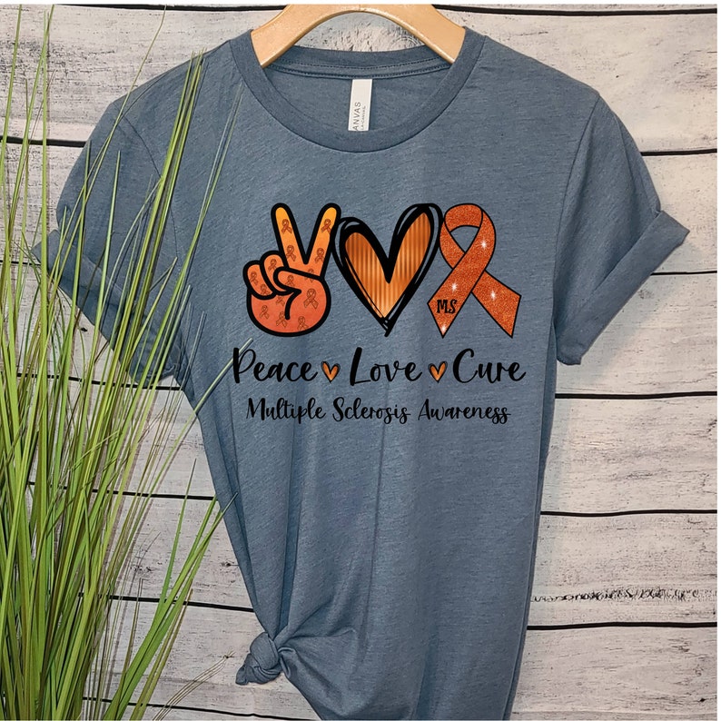 Puede incluir: Una camiseta azul con un signo de paz, un coraz&oacute;n y una cinta naranja con el texto "Peace Love Cure" y "Multiple Sclerosis Awareness".