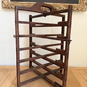 May include: A dark brown, wooden wine rack with multiple angled shelves. The rack has a handle at the top and is designed to hold wine bottles. The rack is made of wood and has a rustic, vintage appearance.