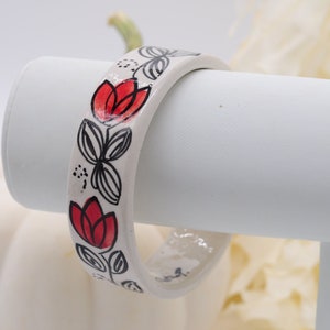 May include: White ceramic bangle bracelet with a black and red floral design. The design features stylized flowers with red petals and black leaves.