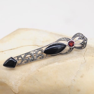 May include: A vintage silver brooch with a geometric design. The brooch features a black onyx stone, a red gemstone, and marcasite accents. The brooch is an elongated shape with a decorative openwork pattern.
