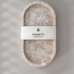 May include: A small oval tray made of white and brown marbled resin. The tray has a smooth, polished finish and is perfect for holding jewelry, keys, or other small items. The tray is labeled "PRIMATTI Small Oval Tray".