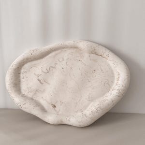May include: A cream-colored, organically shaped tray with a marbled pattern. The tray has a raised edge and a smooth surface. The color palette is neutral, with shades of cream, beige, and brown. The tray is likely made of ceramic or a similar material.