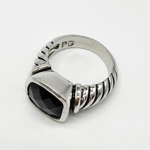 May include: A silver ring with a rectangular black stone. The ring has a ribbed design on the sides and a polished finish. The initials "PD" are engraved on the ring.