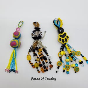 May include: Three beaded jewelry pieces with colorful designs. One features blue, yellow, and pink beads, another has black and amber beads, and the last has a yellow paw print. The text "Peace Of Jewelry" is visible.