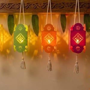 May include: Five hanging lanterns in light blue, yellow, green, orange, and magenta, each featuring intricate cut-out patterns and a central light. Each lantern is suspended by a white cord and finished with a small white tassel at the base.