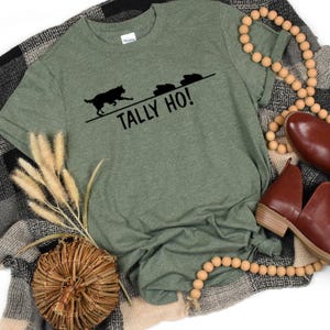 May include: Green t-shirt with the text "TALLY HO!" and a black graphic of a dog chasing three objects. The shirt is displayed on a plaid blanket with brown shoes and wooden beads.