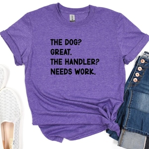 May include: Purple t-shirt with the text "THE DOG? GREAT. THE HANDLER? NEEDS WORK." The shirt is a heathered purple color and has rolled-up sleeves. Part of a pair of blue jeans is visible on the right side of the shirt.