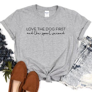 May include: A light gray t-shirt with the text "LOVE THE DOG FIRST and the sport second" in black script. The shirt is styled with brown loafers and denim shorts. The shirt is made of a soft material.