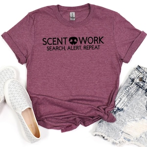 May include: A maroon t-shirt with the words "SCENT WORK" above "SEARCH, ALERT, REPEAT" in black. The shirt is styled with rolled-up sleeves and a knot at the waist. White and gray striped shoes and denim shorts are visible.