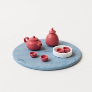 May include: A miniature tea set with a teapot, a small pitcher, and three cups. The set is made of red ceramic and is arranged on a blue circular tray.