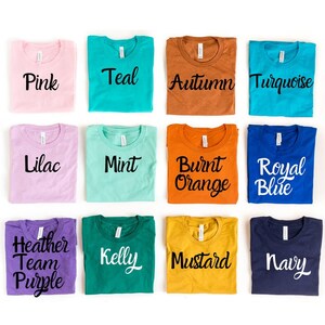 May include: Twelve folded t-shirts in various colors, each with a different color name printed on the front. The colors include pink, teal, autumn, turquoise, lilac, mint, burnt orange, royal blue, heather team purple, kelly, mustard, and navy.