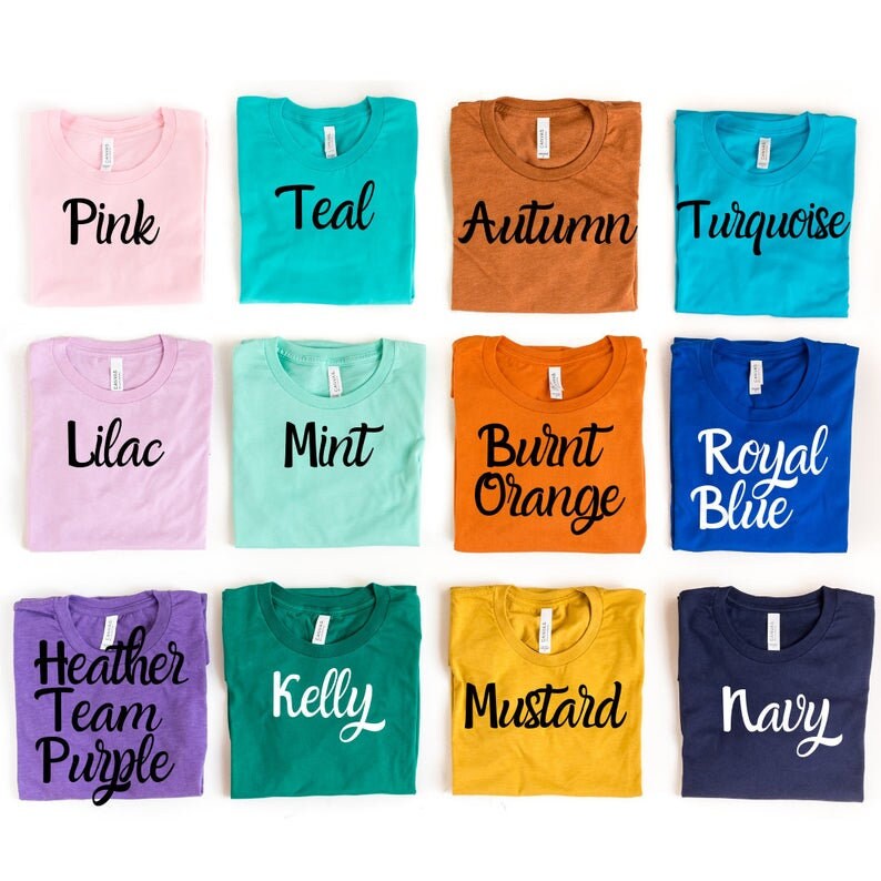 May include: Twelve folded t-shirts in various colors, each with a different color name printed on the front. The colors include pink, teal, autumn, turquoise, lilac, mint, burnt orange, royal blue, heather team purple, kelly, mustard, and navy.