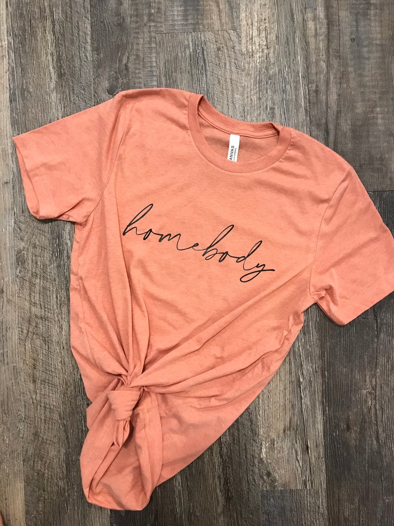 May include: A peach colored t-shirt with the word "homebody" written in black cursive lettering.