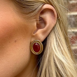 Peut inclure: Boucles d'oreilles à clous dorées avec une pierre centrale ovale rouge foncé. Les boucles d'oreilles ont une bordure dorée à double couche. L'image montre un gros plan des boucles d'oreilles sur une oreille, avec des cheveux blonds en arrière-plan.