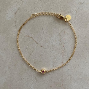 Op de afbeelding: Een gouden kettingarmband met een klein rood edelsteentje in het midden.