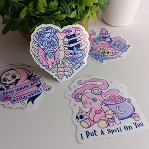 May include: Four pastel pink and purple aesthetic stickers with spooky and cute designs. The stickers feature a skeleton heart with a bat, a skeleton in a pink hooded robe, a teddy bear with a cauldron, and a potion bottle with a skull and crossbones. The text on the stickers reads "Dead Inside", "Dead Inside But Still Cute", "I Put A Spell On You", and "Don't Open Me".