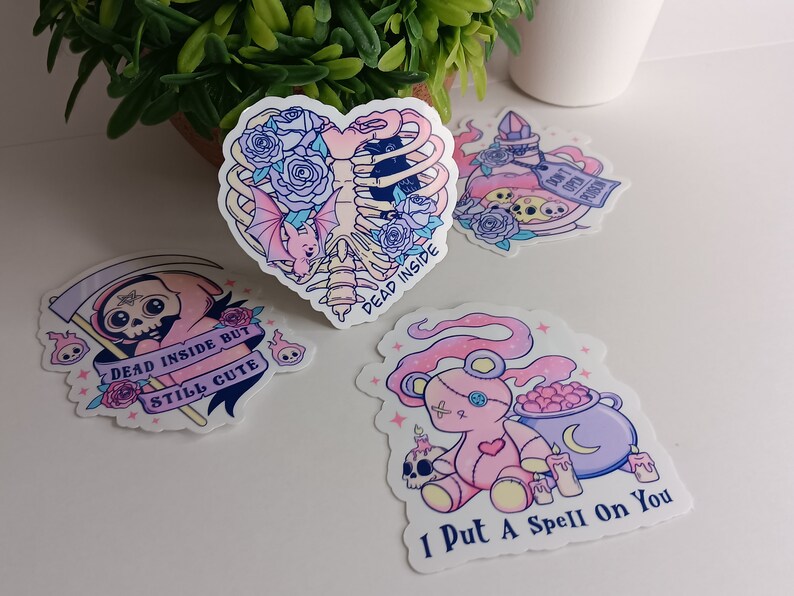 May include: Four pastel pink and purple aesthetic stickers with spooky and cute designs. The stickers feature a skeleton heart with a bat, a skeleton in a pink hooded robe, a teddy bear with a cauldron, and a potion bottle with a skull and crossbones. The text on the stickers reads "Dead Inside", "Dead Inside But Still Cute", "I Put A Spell On You", and "Don't Open Me".