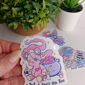 May include: A pink and white sticker with a cartoon pink teddy bear holding a cauldron with a crescent moon. The teddy bear has a stitched heart and an X on its chest. The sticker says "I Put A Spell On You".