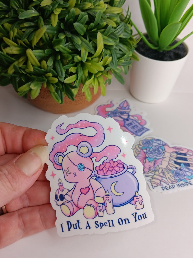 May include: A pink and white sticker with a cartoon pink teddy bear holding a cauldron with a crescent moon. The teddy bear has a stitched heart and an X on its chest. The sticker says "I Put A Spell On You".