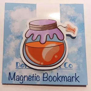 May include: A magnetic bookmark featuring a cartoon illustration of a honey jar with a purple lid and orange honey. The bookmark has the text "Magnetic Bookmark" and a small tag that says "Honey". The background is light blue with a cloud design.