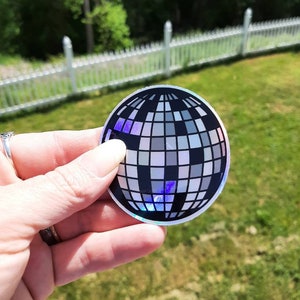 May include: A silver holographic sticker of a disco ball. The sticker is round and has a black and white design.