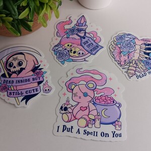 May include: Four pink and purple stickers with gothic and cute designs. The stickers feature a skeleton with a bat, a potion bottle with a skull, a pink teddy bear with a cauldron, and a pink grim reaper with a scythe. The text on the stickers reads "Dead Inside But Still Cute", "Don't Open Poison", "I Put A Spell On You", and "Dead Inside".