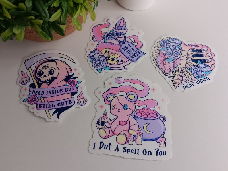 May include: Four pink and purple stickers with gothic and cute designs. The stickers feature a skeleton with a bat, a potion bottle with a skull, a pink teddy bear with a cauldron, and a pink grim reaper with a scythe. The text on the stickers reads "Dead Inside But Still Cute", "Don't Open Poison", "I Put A Spell On You", and "Dead Inside".