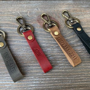 May include: Four leather keychains in gray, red, tan, and black. Each keychain has a metal clasp and a leather strap with engraved text. The gray keychain says "DRIVE SAFE", the red says "ADW", the tan has a phone number, and the black says "NICK".