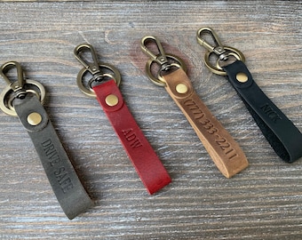 May include: Four leather keychains in gray, red, tan, and black. Each keychain has a metal clasp and a leather strap with engraved text. The gray keychain says "DRIVE SAFE", the red says "ADW", the tan has a phone number, and the black says "NICK".