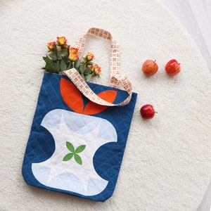 May include: A blue quilted tote bag with a white apple design and orange leaves. The bag has a white and orange patterned strap. The bag is on a white fuzzy surface with red apples scattered around it.
