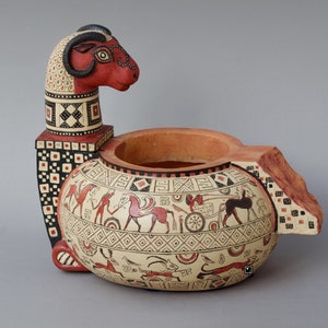 Può includere: Un vaso in ceramica rosso e bianco a forma di ariete con motivi geometrici e una scena di cavalli e carri dipinti sul lato.