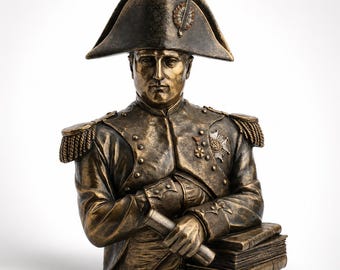 May include: A bronze-toned bust of Napoleon Bonaparte wearing a bicorne hat and a military uniform with epaulets, medals, and a high collar. The figure holds a rolled document in one hand and rests the other arm on a stack of three thick books.