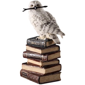 May include: A white and gray owl figurine with a twig in its beak, perched atop a stack of antique-style books. The books have titles like "Witchcraft" and "The Ars Notoria" in gold lettering, creating a mystical theme.
