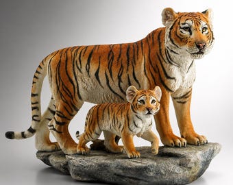 May include: A detailed figurine of a mother tiger and her cub. The adult tiger stands protectively over the cub, both with orange and black striped coats. They are posed on a stone-like base, showcasing the intricate details of the sculpture.