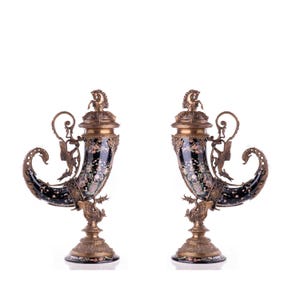 May include: Pair of ornate, black and gold decorative cornucopia vases. Each vase features a curved horn shape with floral designs, bronze accents, and a detailed base. The vases are likely for display.