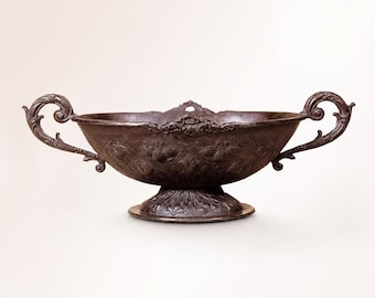May include: A detailed, antique-style metal bowl with ornate handles and a decorative base. The bowl has a dark, aged finish and features embossed floral patterns. The handles are scrolled, adding to the vintage aesthetic. The bowl is centered on a plain background.