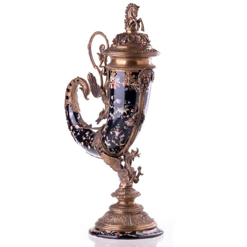 May include: An ornate, black and gold decorative horn-shaped vessel. The vessel features intricate floral and dragon designs, with a horse figurine on the lid. The base and accents are gold-toned, creating a luxurious aesthetic. A unique decorative piece.